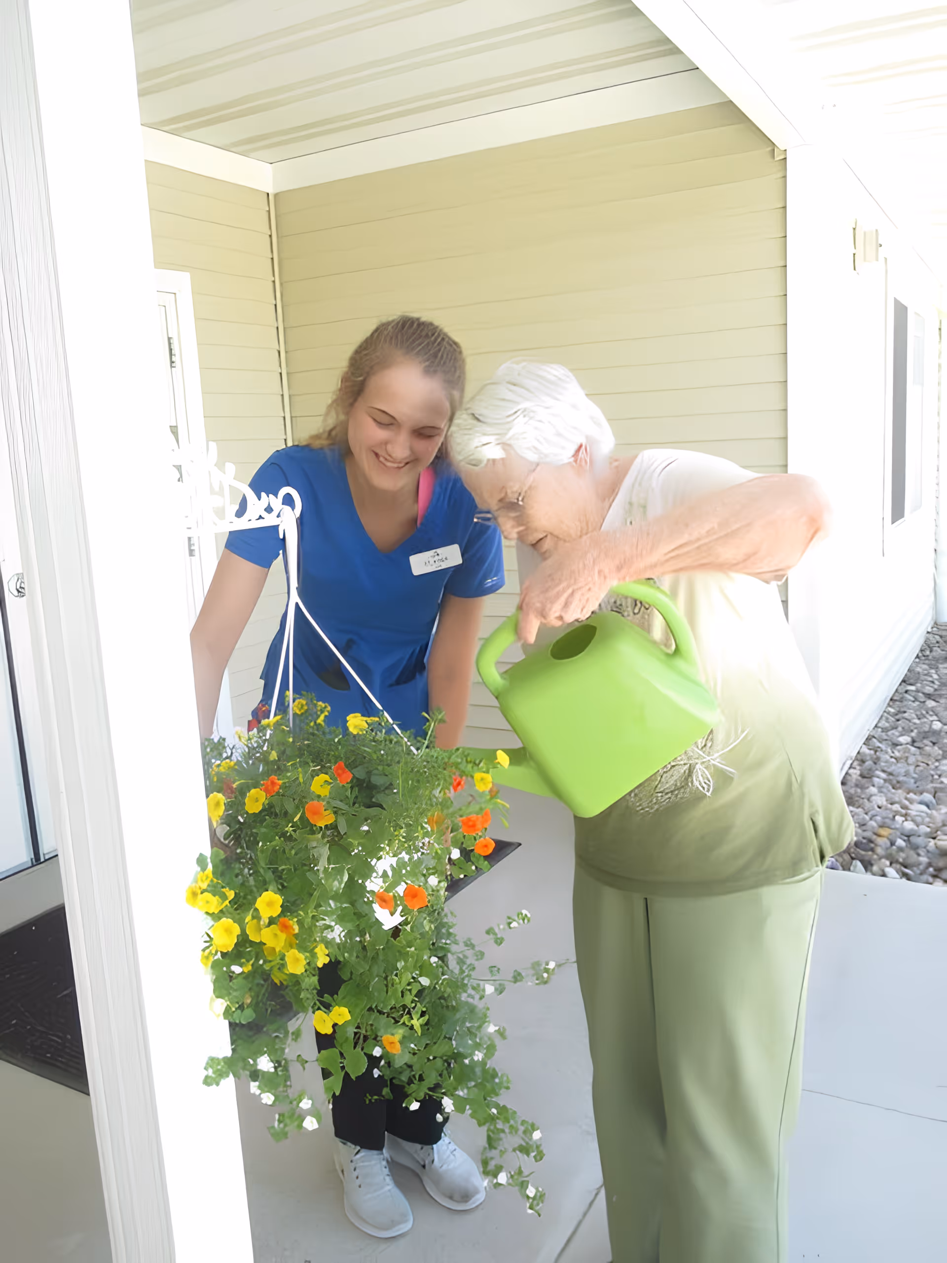 An elderly woman watering a hanging flower basket with a green watering can while a younger woman in a blue shirt smiles and watches her on a covered porch.
