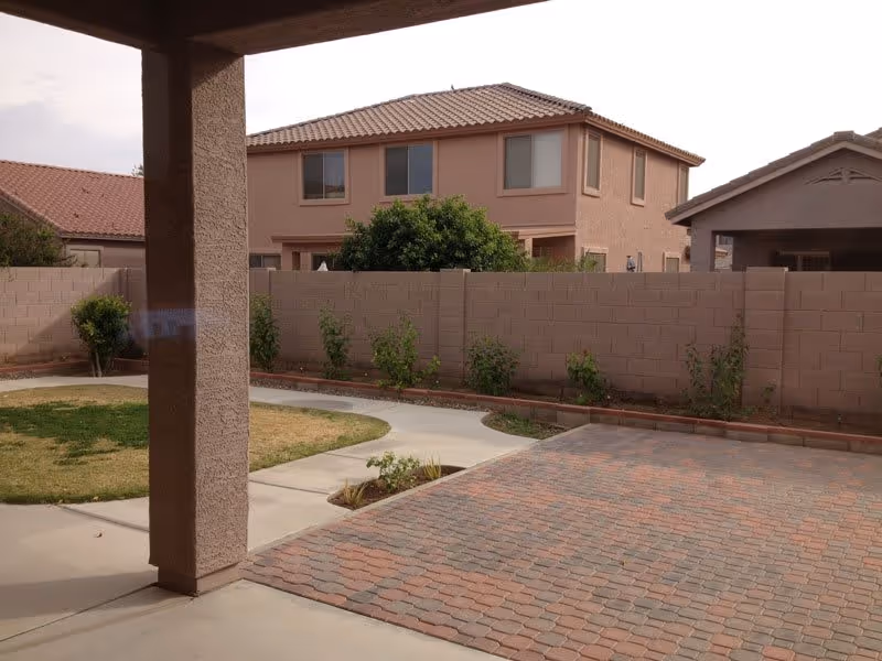 View from a covered patio over a brick-paved patio and small lawn toward a cinderblock backyard wall and a two-story neighboring house.