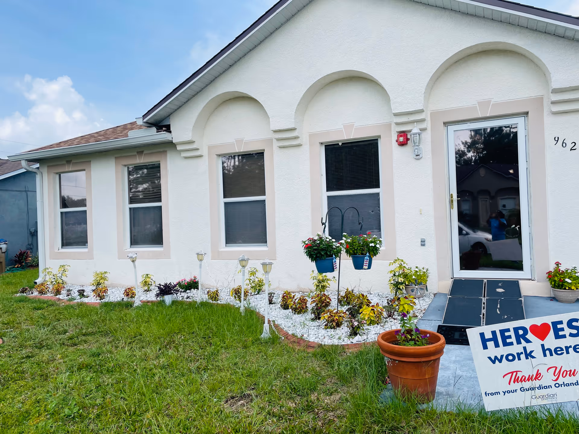 Exterior view of a white building with three windows and a glass door. There is a small garden with flowers and plants in front of the building, along with a sign that reads 'HEROES work here Thank You from your Guardian Orlando'. The sky is partly cloudy.
