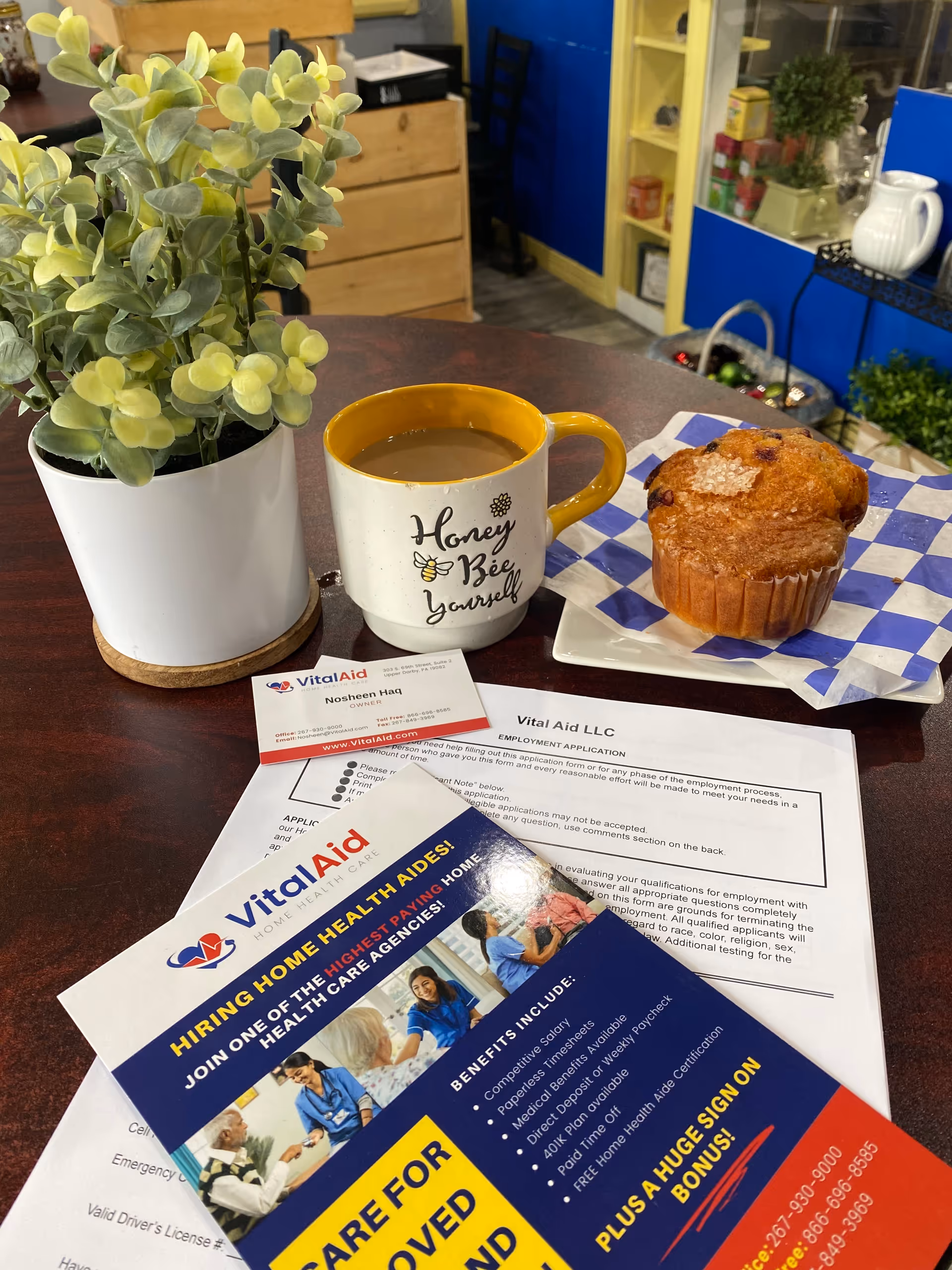 A desk scene with a potted plant, mug, muffin, and Vital Aid brochures.