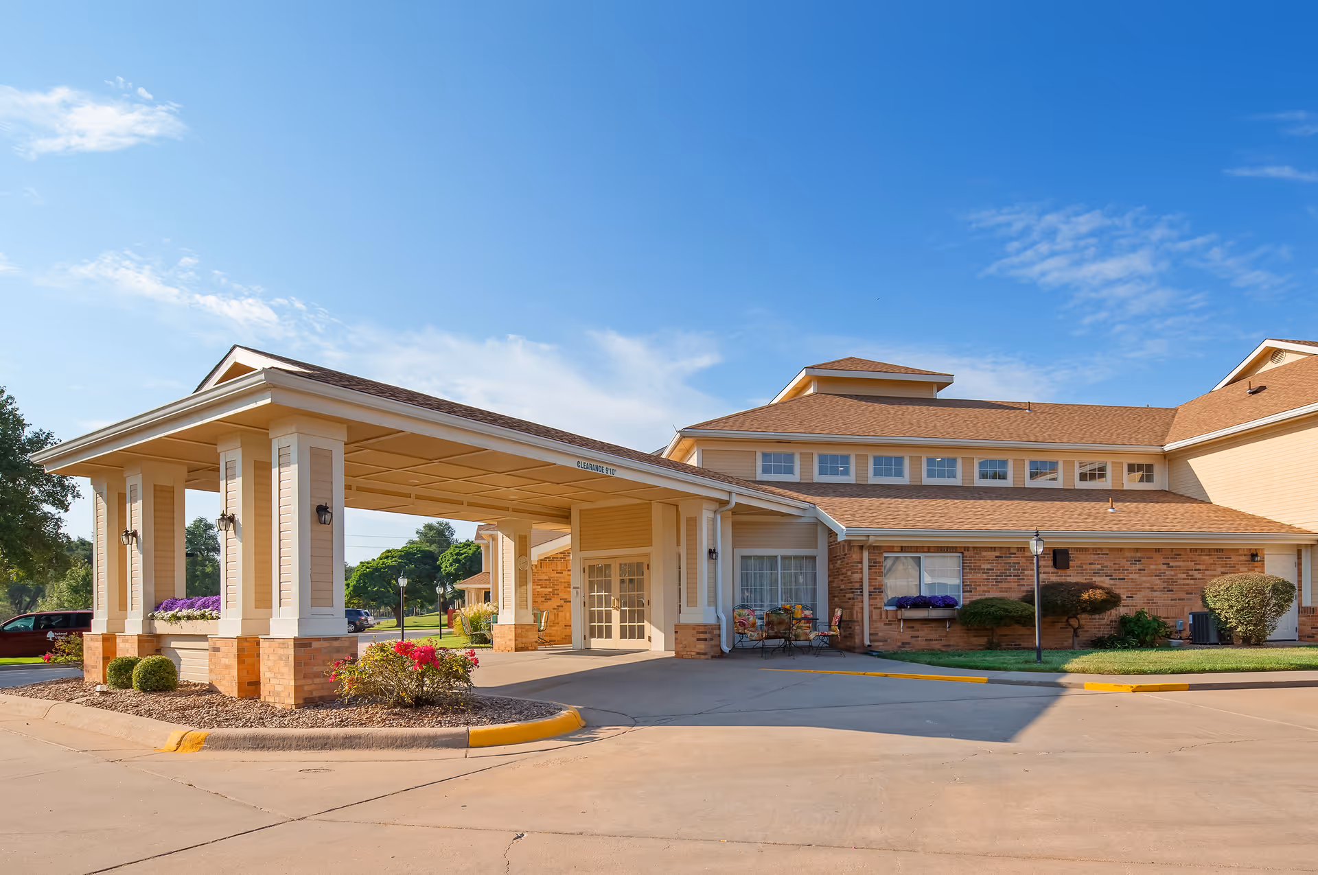 Exterior view of Parkwood Village senior living facility showing the main entrance with a covered drop-off area, brick and beige siding, landscaped bushes and flowers, and a clear blue sky.
