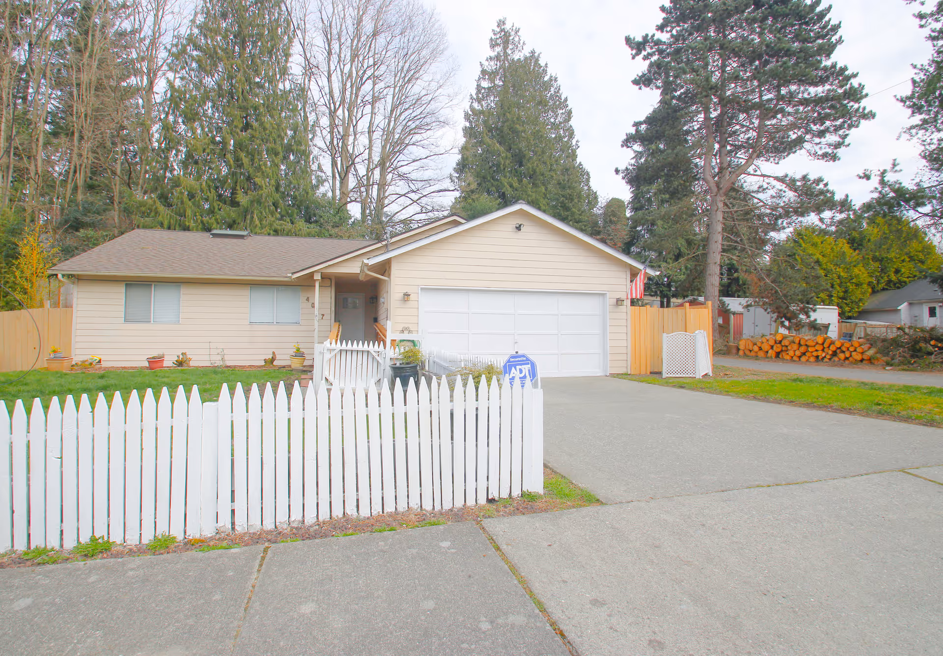 A single-story beige house with a white picket fence and driveway, surrounded by tall trees.