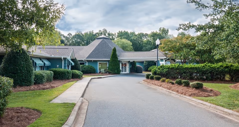 Exterior view of a single-story senior living facility building with a paved driveway leading to the entrance. The building is surrounded by well-maintained bushes, trees, and green lawns under a partly cloudy sky.