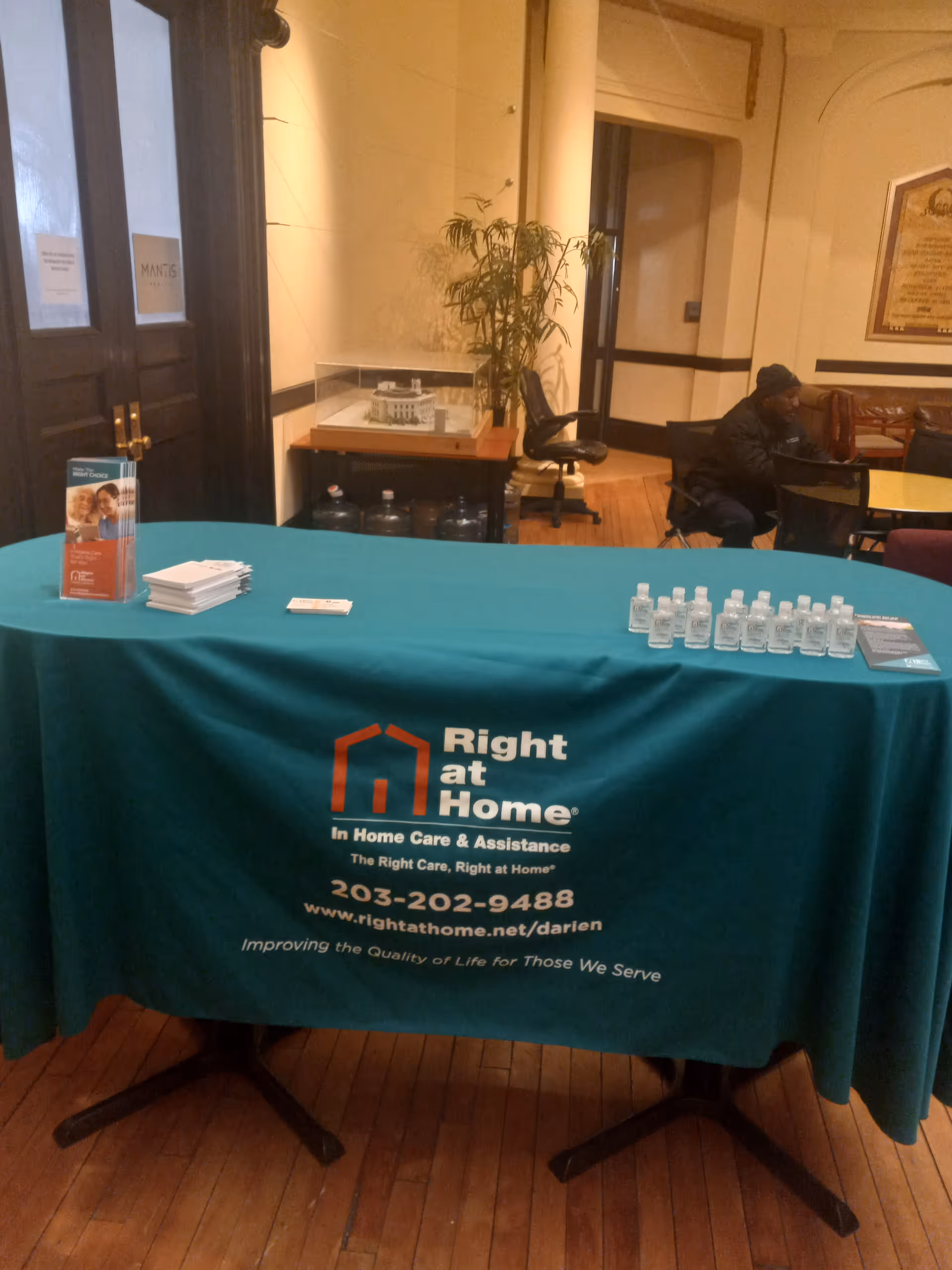 Reception table with teal cloth, brochures, sanitizer, and a person seated in the background.