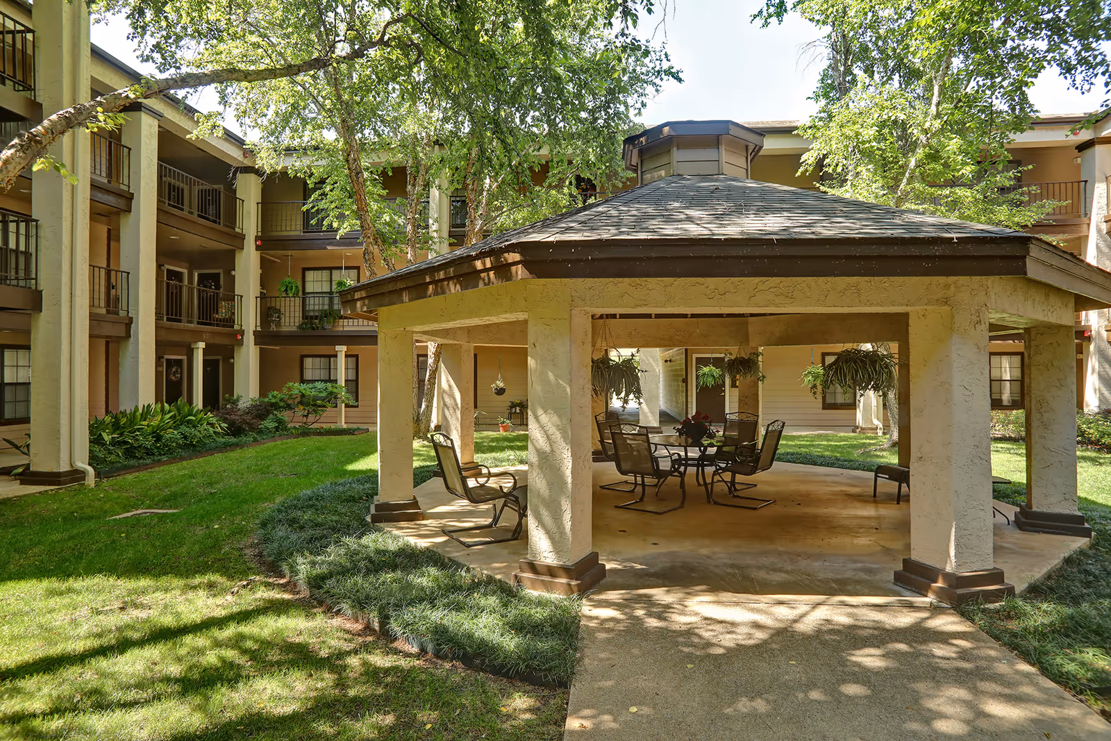 Outdoor gazebo with a hexagonal roof and several chairs and a table underneath, surrounded by green grass and trees, with a multi-story residential building in the background.