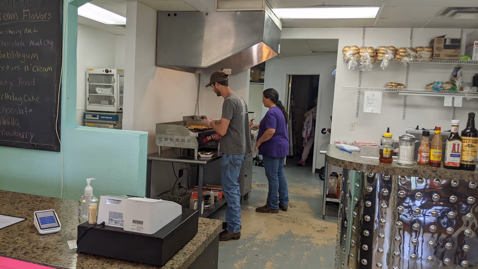 Staff members cook at a grill in a bustling senior living facility kitchen.