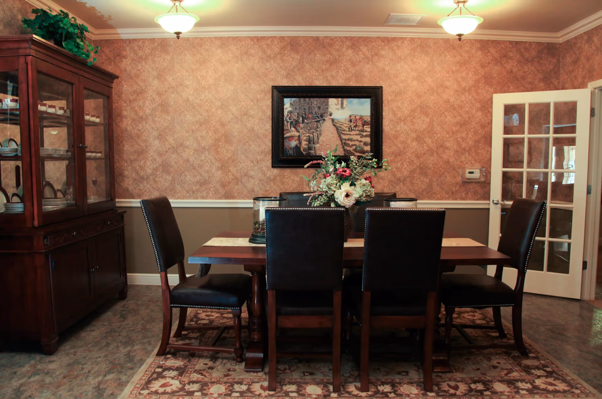 A formal dining room with a wooden dining table surrounded by six dark leather chairs. The table has a floral centerpiece and two candle holders. There is a wooden china cabinet on the left filled with dishes, a framed painting on the wall, and a glass-paneled door on the right. The room has patterned wallpaper, two ceiling lights, and a decorative rug under the table.