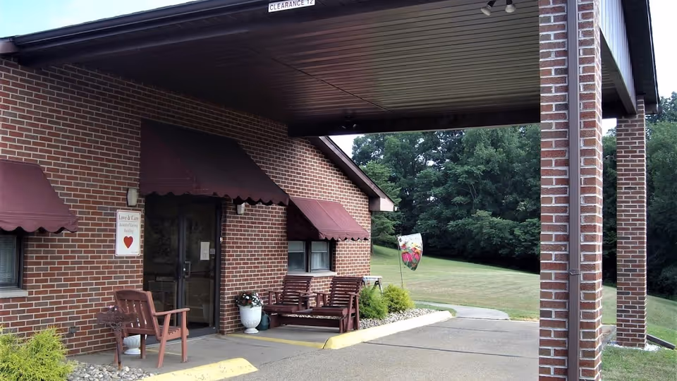 Entrance of a brick building with a covered driveway. There are maroon awnings over the windows and door. Two wooden benches and a chair are placed near the entrance, along with potted plants. A sign next to the door reads 'Love & Care Assisted Living Family' with a red heart. The background shows a grassy area and trees.