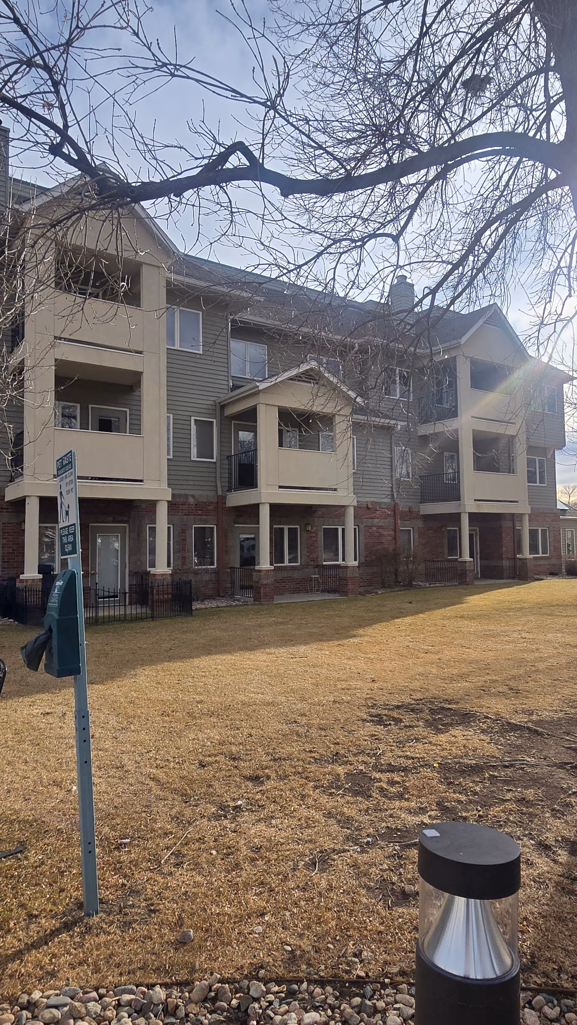Three-story senior living building exterior with balconies, leafless trees, and a grassy yard.
