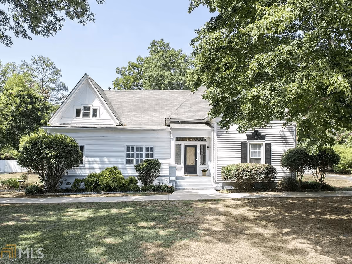 White single-story house front with a central doorway, shrubs, and a shaded lawn under large trees.
