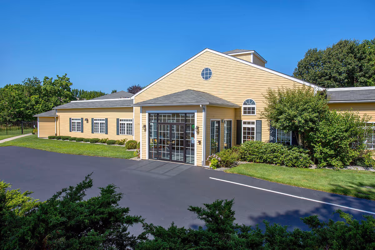Exterior view of a single-story yellow building with white trim and multiple windows, surrounded by green bushes and trees under a clear blue sky. The building has a large glass entrance with a small roof overhang and a paved driveway in front.
