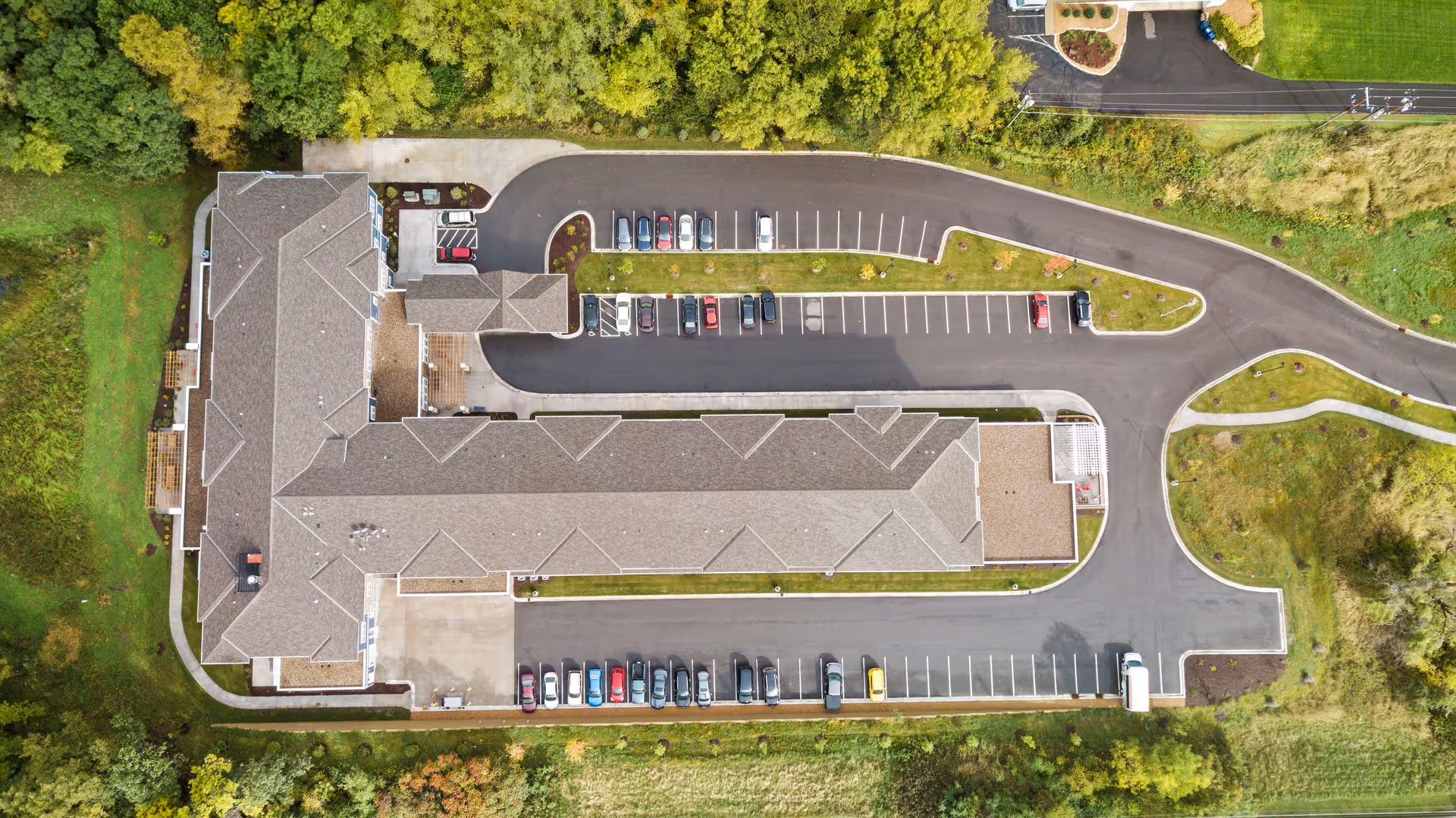 Aerial view of Harrison Bay Senior Living facility showing a large building with a gray roof surrounded by parking lots with several cars parked. The building is bordered by green trees and grass areas.