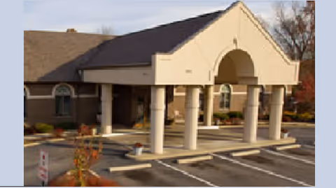 Exterior view of a single-story building with a covered entrance supported by columns, surrounded by a parking lot with marked spaces and some landscaping.