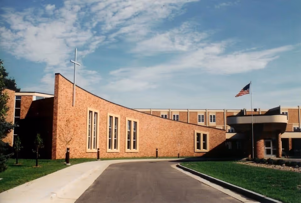 Brick senior living facility with curved entrance, cross sculpture, flagpole, and blue sky.