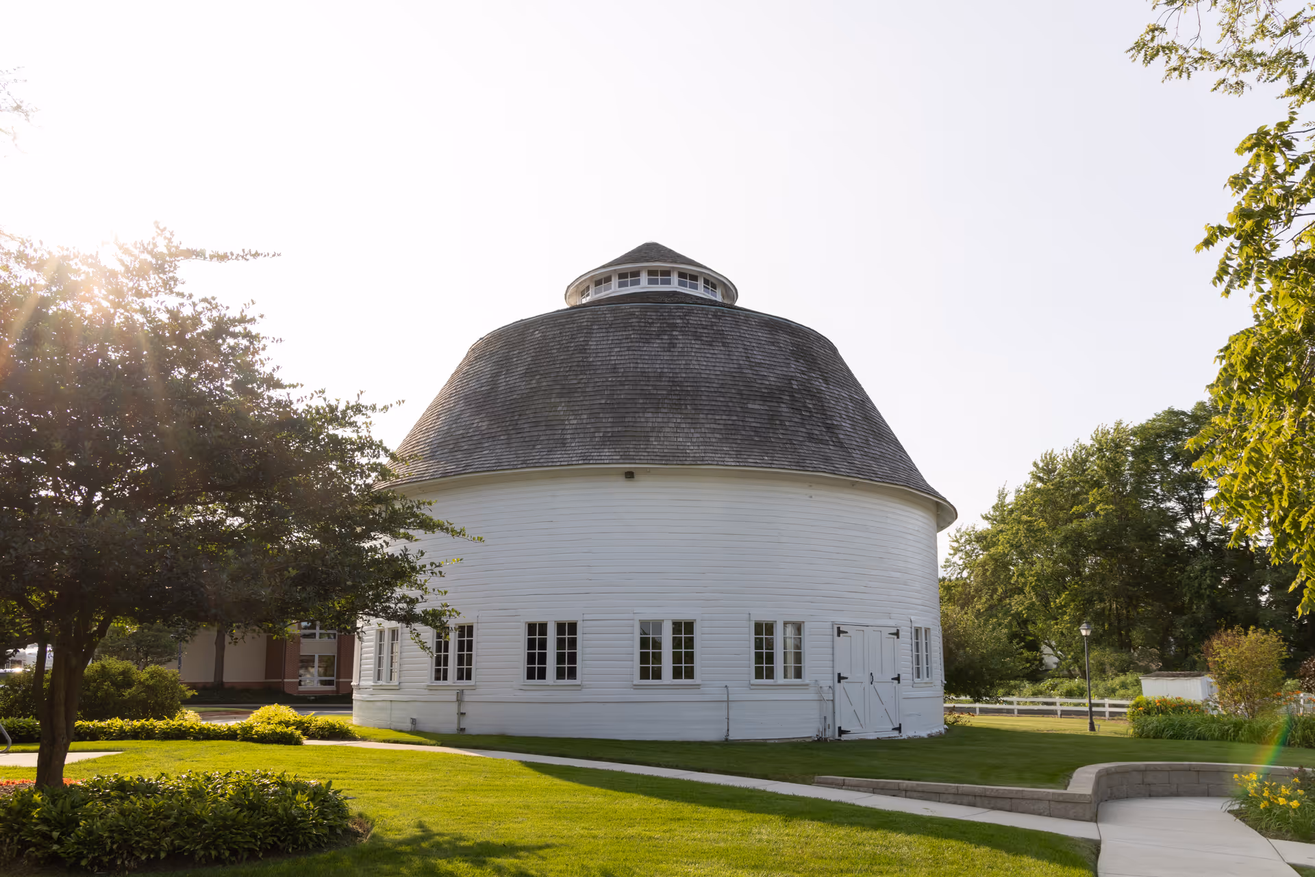 A round white building with a domed shingled roof on a manicured lawn with trees and walkways in bright sunlight.