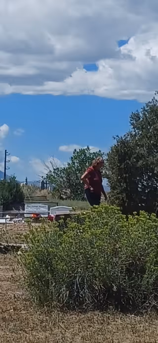 A person stands outdoors near shrubs with parked cars and blue sky.
