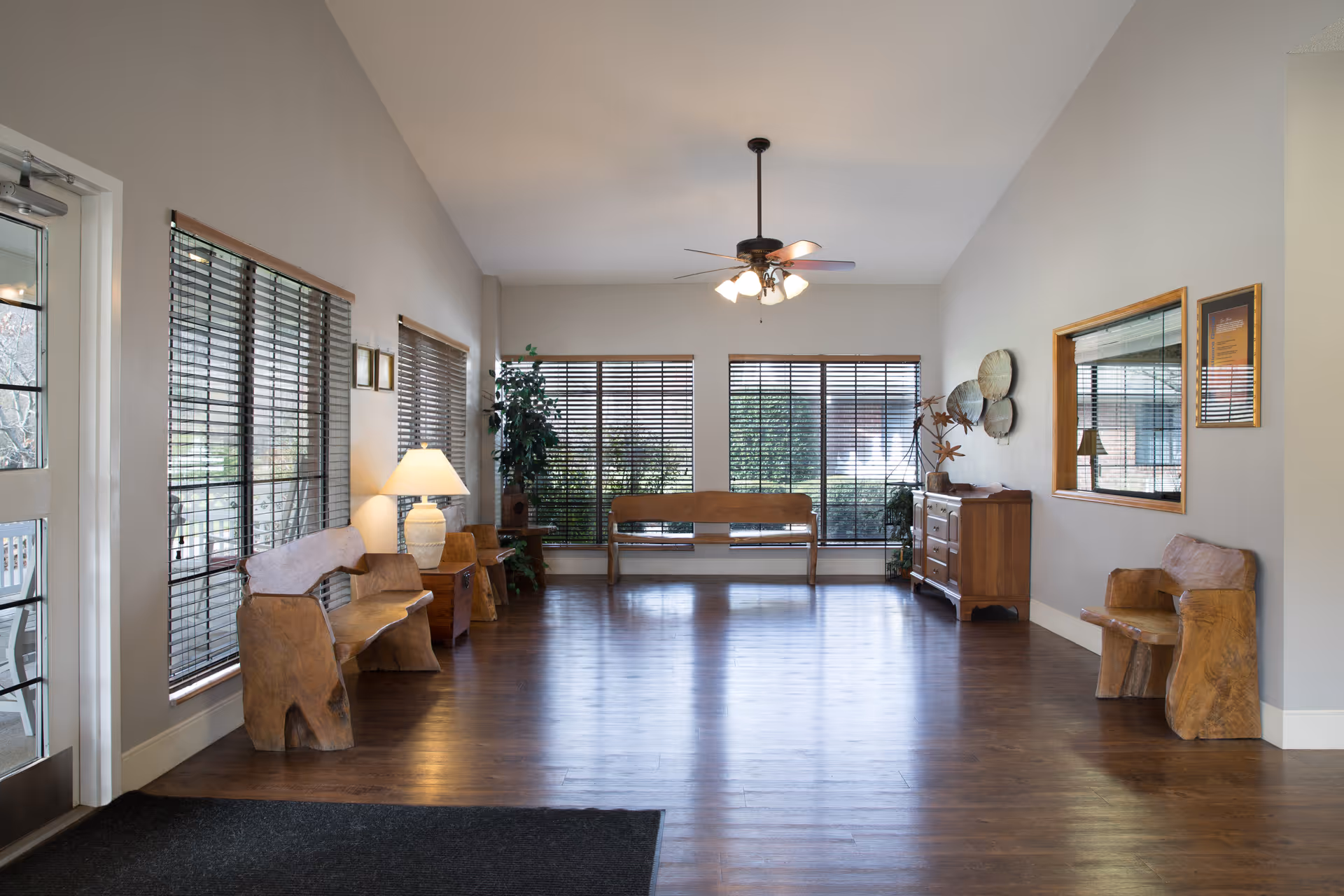 Bright lounge with wooden benches, tables, windows, and a ceiling fan in a senior living facility.