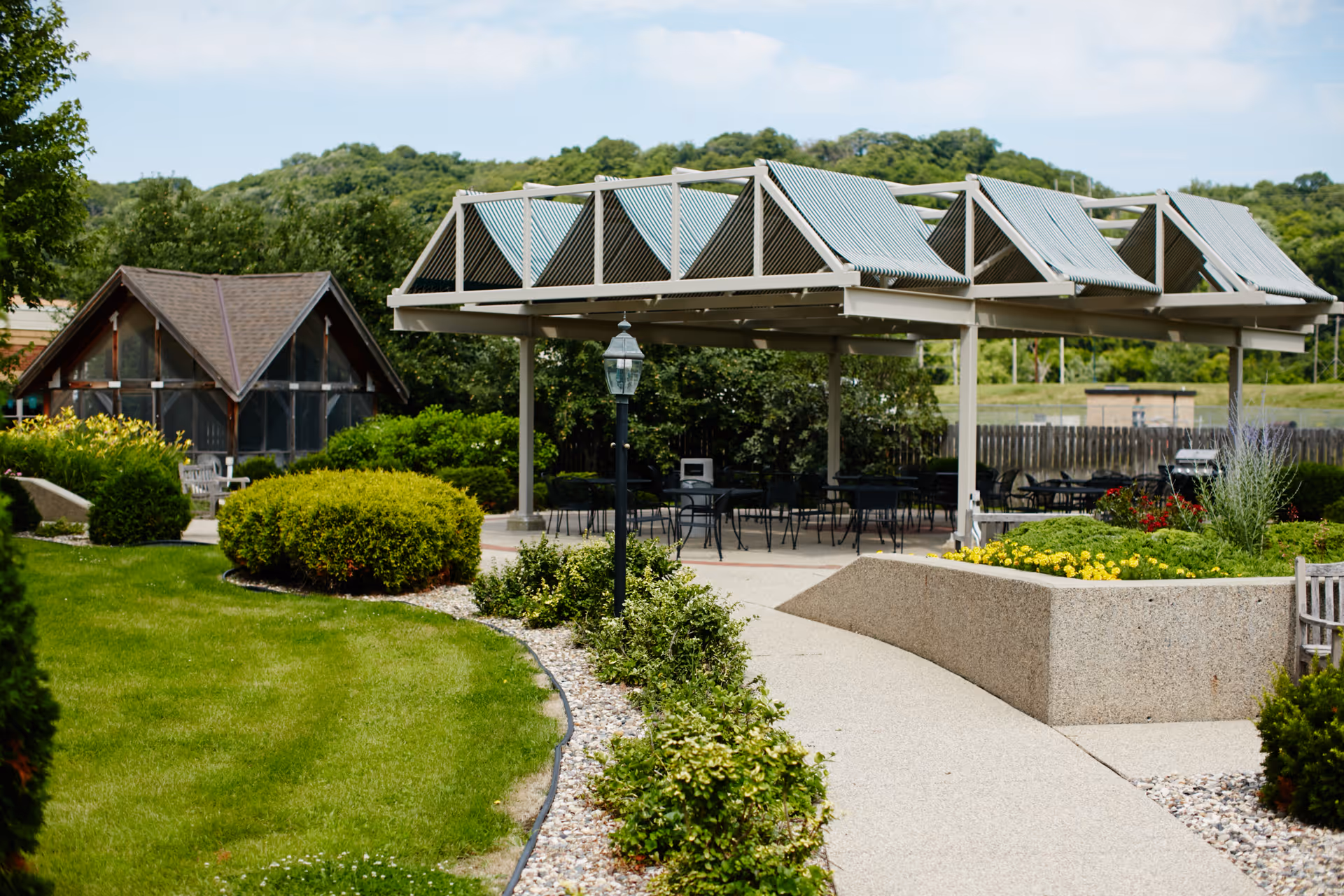 Outdoor seating area with a modern pergola structure providing shade over several tables and chairs. Surrounding the area are well-maintained green bushes, flower beds, and a small building with a peaked roof in the background. The scene is set in a landscaped garden with a paved walkway and a lamppost.