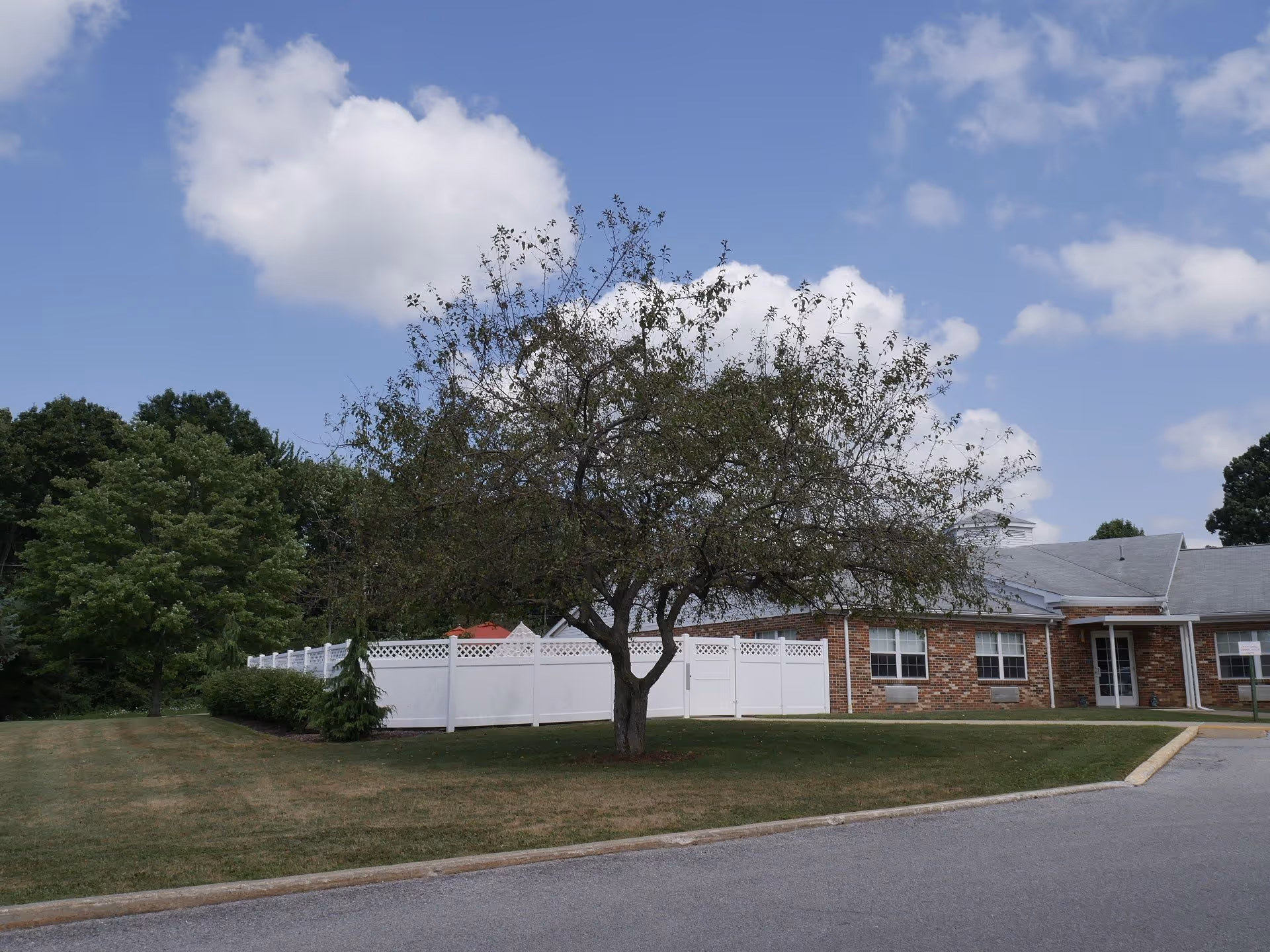 Exterior view of Ridgeview Healthcare and Rehabilitation Center showing a brick building with white trim, a white fence, a tree in the foreground, and a partly cloudy sky.