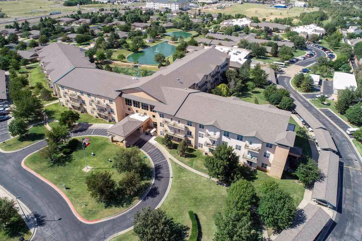 Aerial view of The Craig Senior Living facility showing a large, multi-wing building surrounded by green lawns, trees, and a circular driveway with a flagpole. There are parking areas and a pond visible in the background along with other buildings and roads.