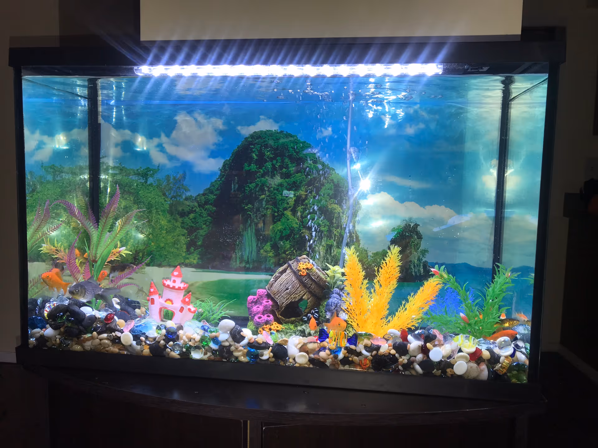 Colorful aquarium with gravel, artificial plants, a small castle, and rocks indoors on a dark stand.