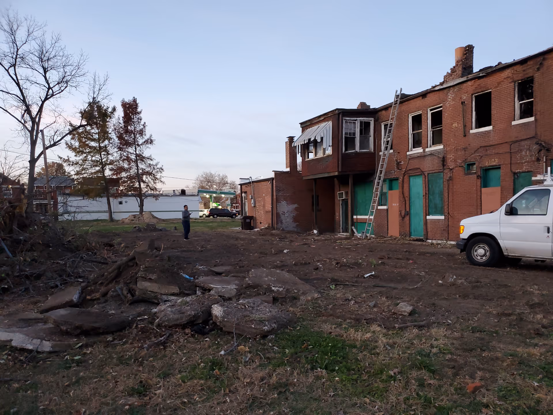 Dilapidated brick building exterior with broken windows, a ladder, and a white van amid rubble.