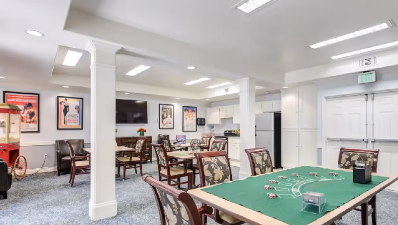 A bright communal recreation room in a senior facility with a poker table, chairs, and kitchenette.