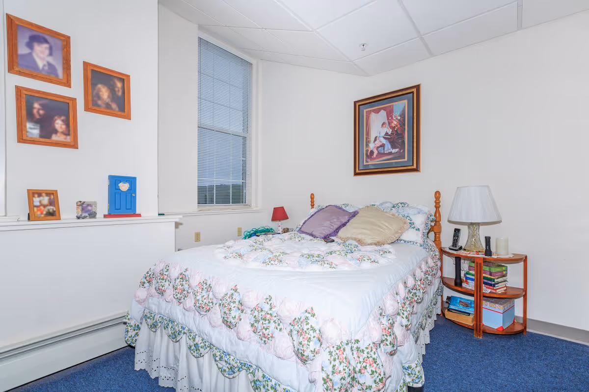 Small bedroom with a floral quilted bed, bedside table with lamp and books, a window, and framed pictures on the walls.