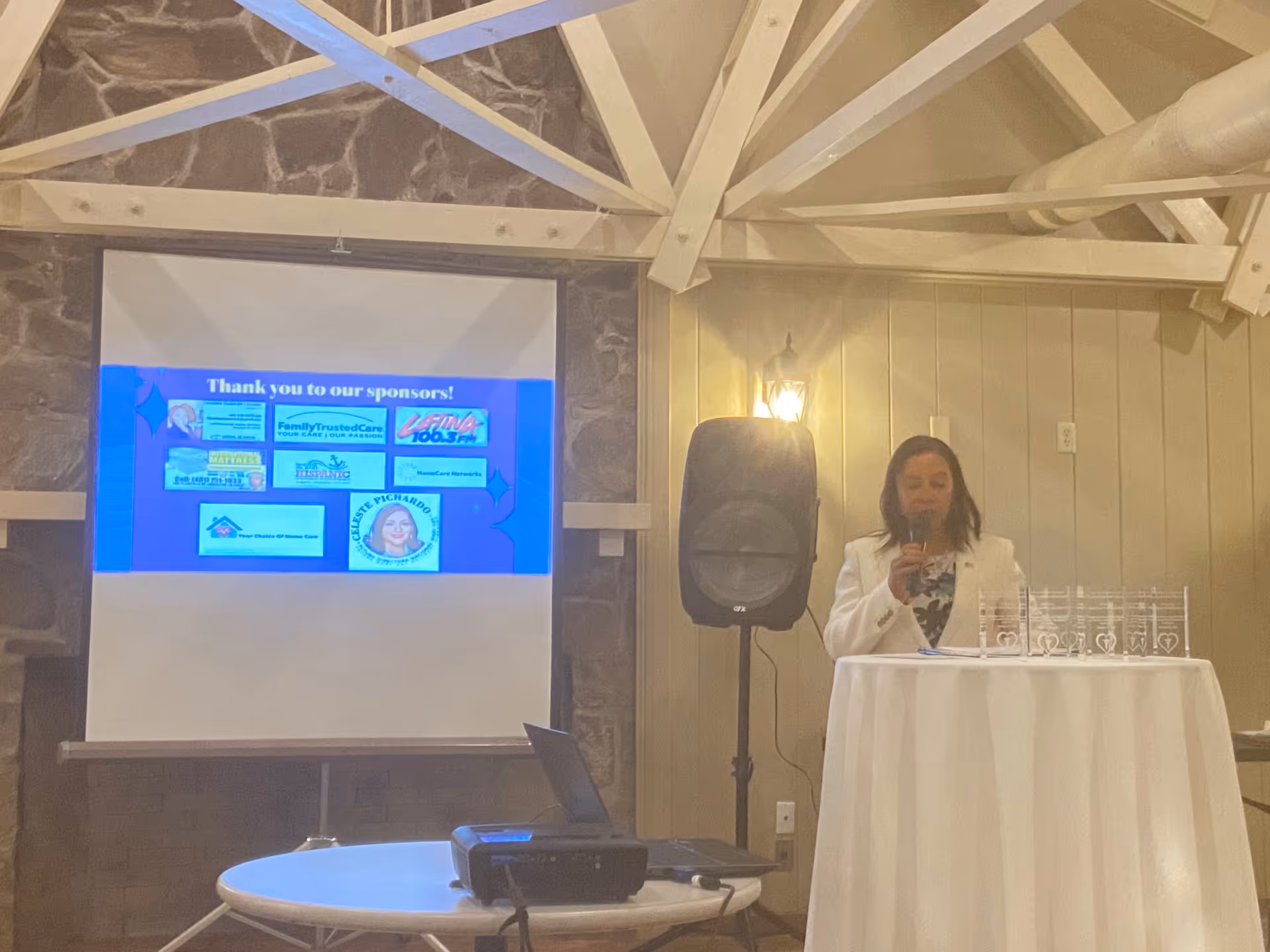 A female presenter speaks at an indoor senior living event with projector screen and awards.