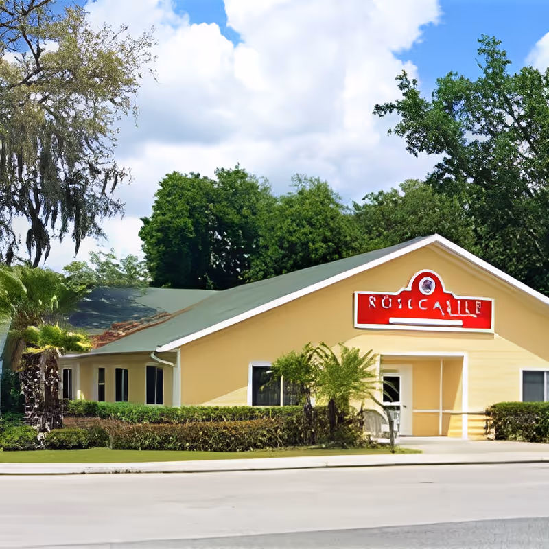 Front exterior of a yellow single-story building with a green roof, a red sign above the entrance, and trees in the background.