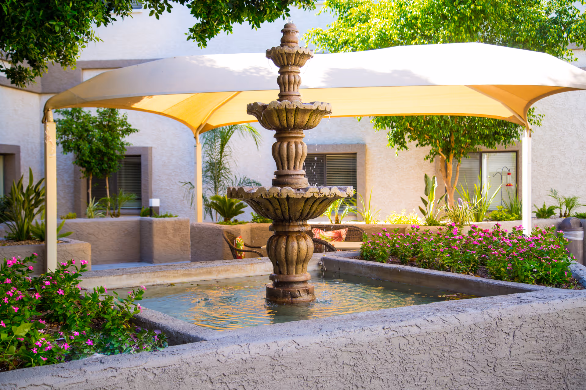 Outdoor courtyard area with a three-tiered stone fountain in the center surrounded by a square water basin. There are flowering plants and greenery around the fountain, with a beige canopy providing shade. The background shows a building wall with windows and more plants.