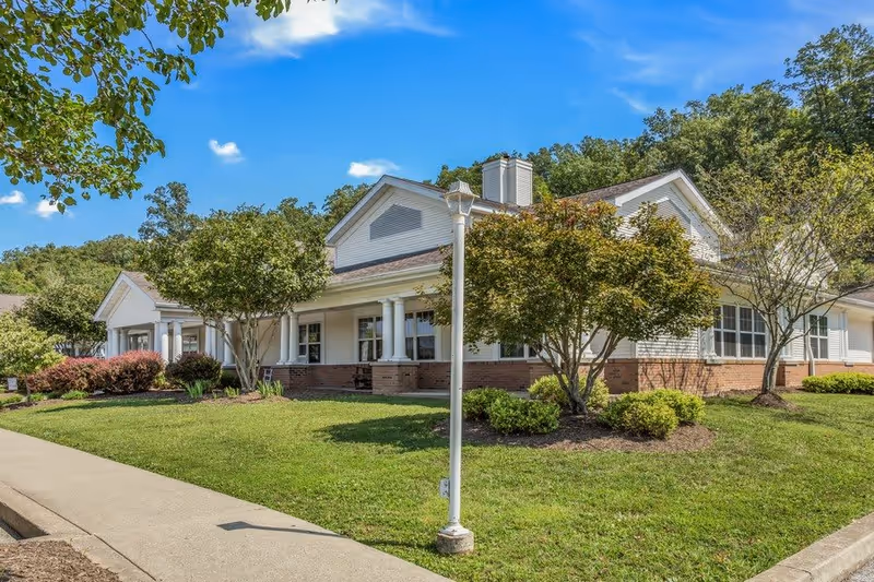 Exterior view of a single-story senior living facility building with white siding and brick accents, surrounded by green grass, trees, and shrubs under a blue sky with a few clouds. A sidewalk and a white lamp post are visible in the foreground.