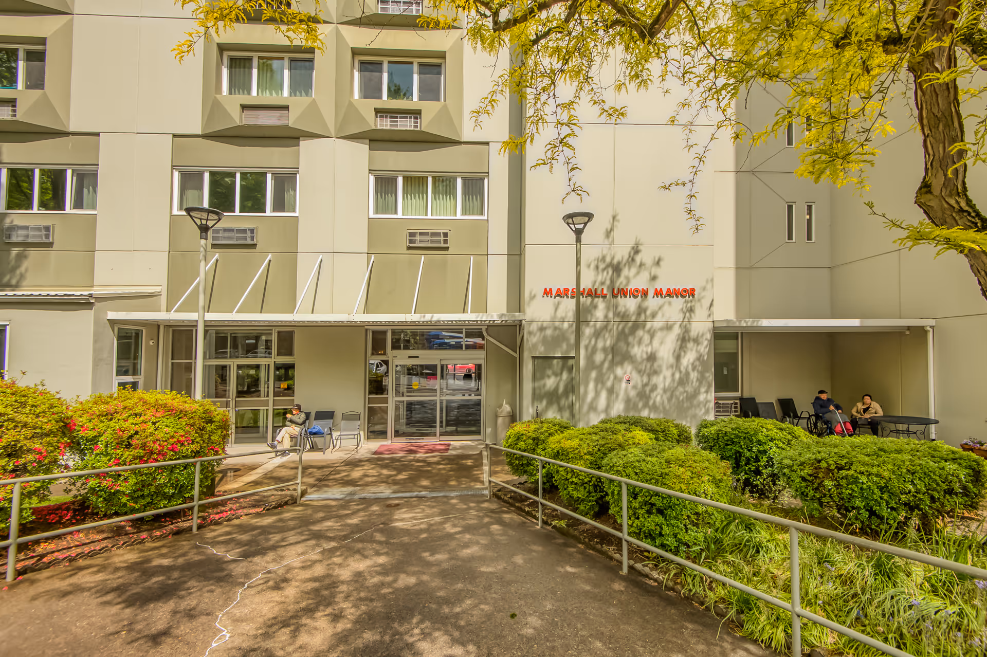 Exterior view of Marshall Union Manor building entrance with a walkway leading to glass doors. There are bushes and trees around the entrance, and two people sitting on chairs near the entrance on the right side.
