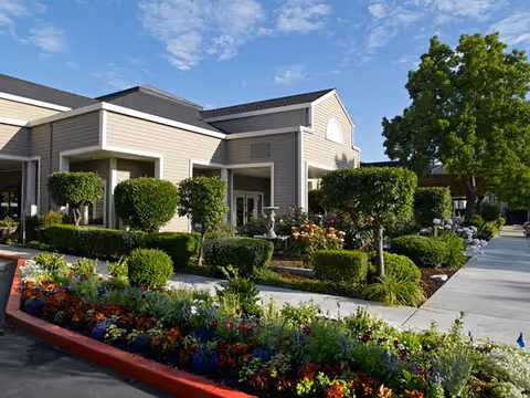 Modern senior living facility exterior with manicured shrubs, colorful flower beds, and a paved entrance.