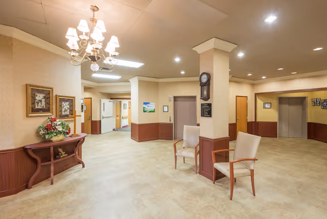 A spacious, well-lit interior hallway area in a senior care facility with beige walls and flooring. There is a wooden table with a floral arrangement and framed pictures on the left wall. Two beige chairs with wooden armrests are placed near a column with a clock and signage. Multiple doors and an elevator are visible in the background.