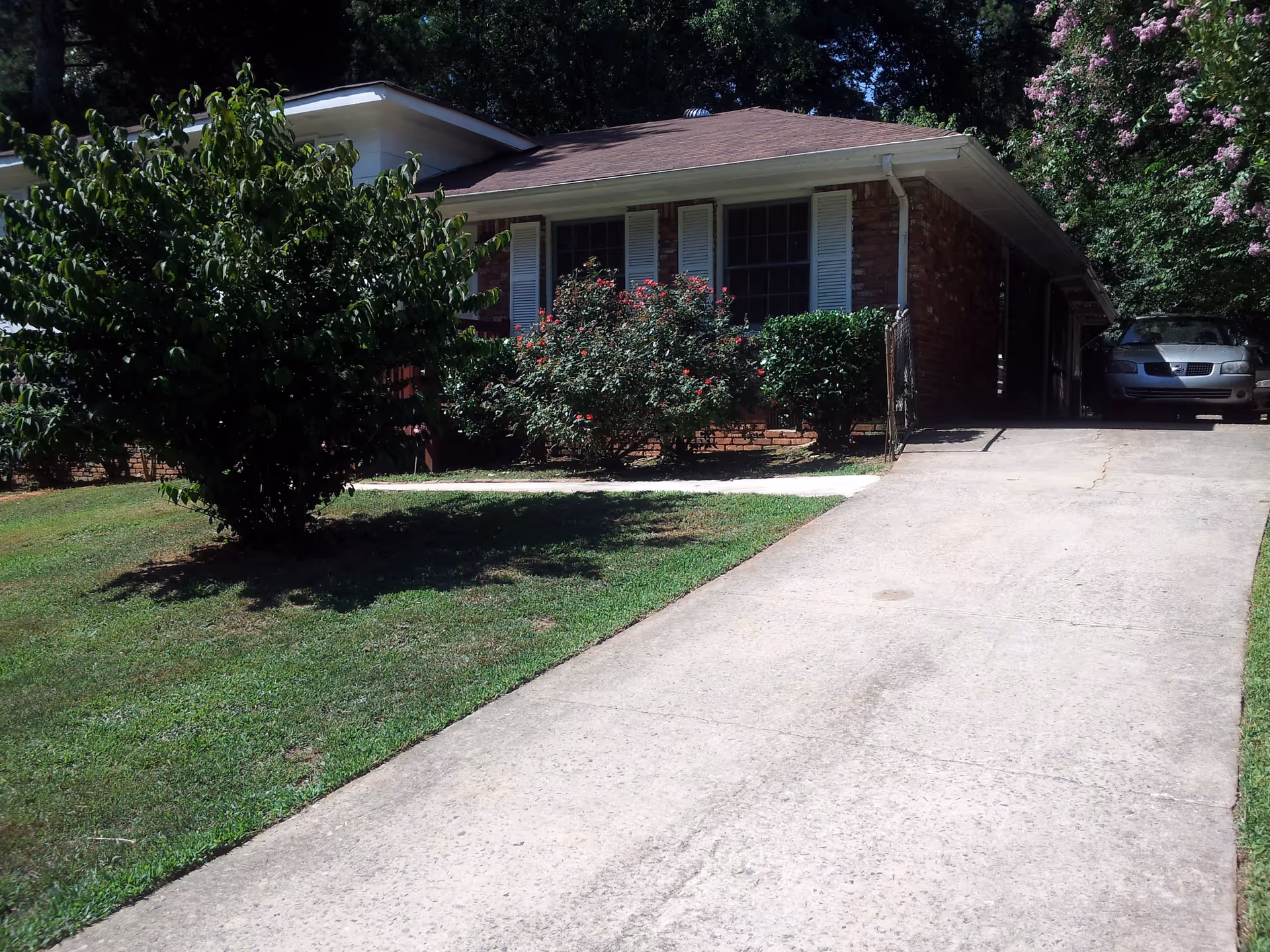 A single-story brick house with white shutters on the windows, surrounded by green bushes and trees. There is a concrete driveway leading up to a carport with a parked car. The lawn is well-maintained with green grass.
