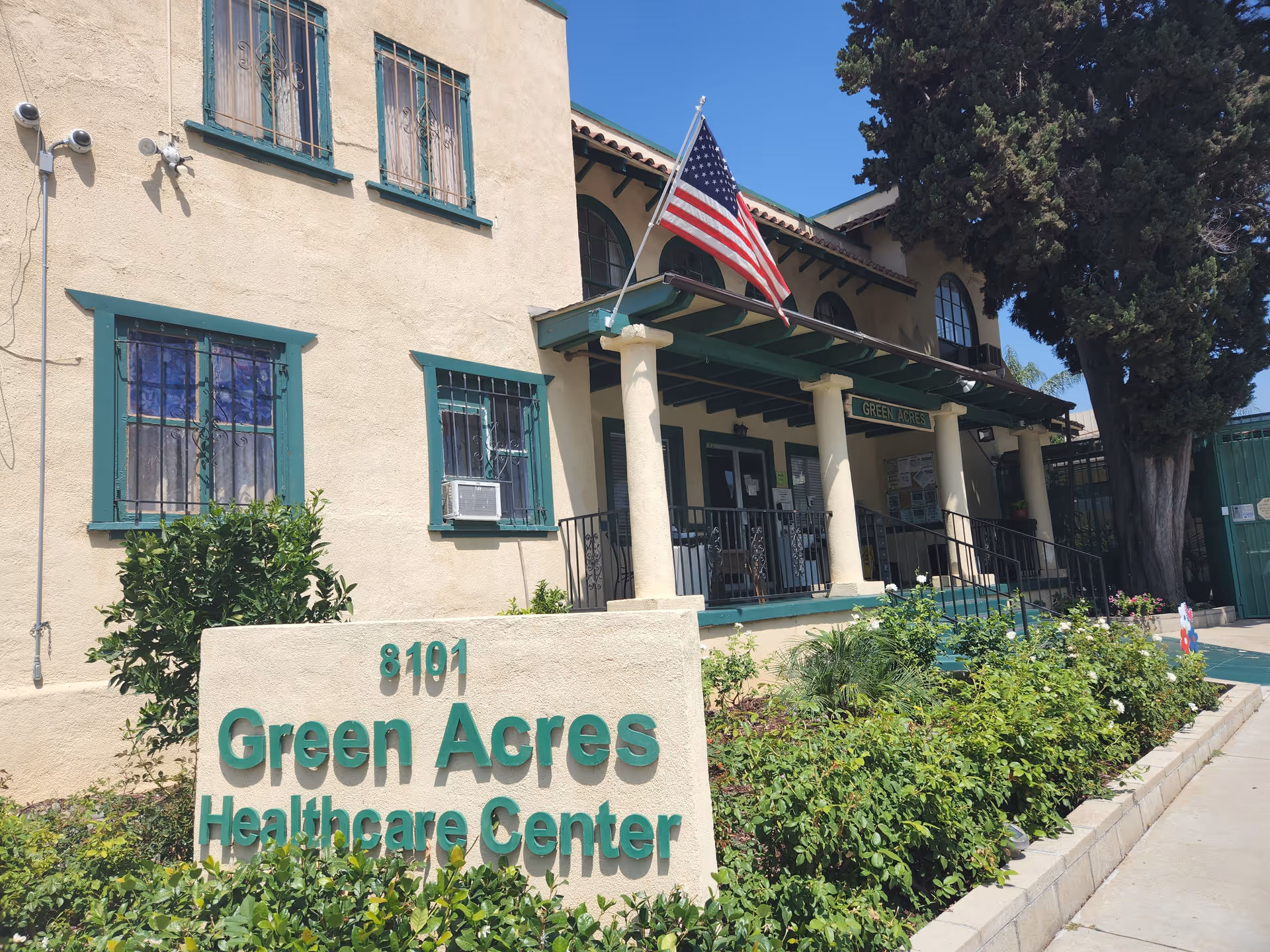 Cream building with green trim, porch, American flag, and a sign reading Green Acres Healthcare Center.