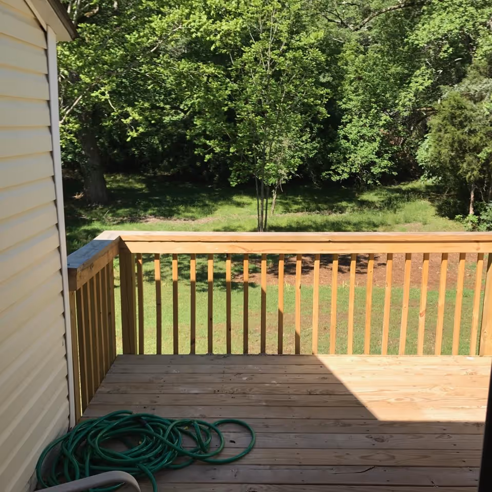 Wooden deck with railing and a green hose overlooking a grassy yard and trees.