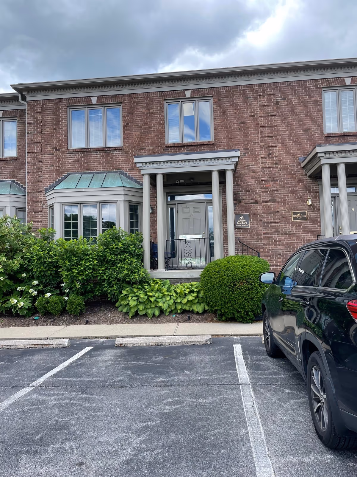 Brick senior living building exterior with columns, entry door, landscaping, and a parked car.