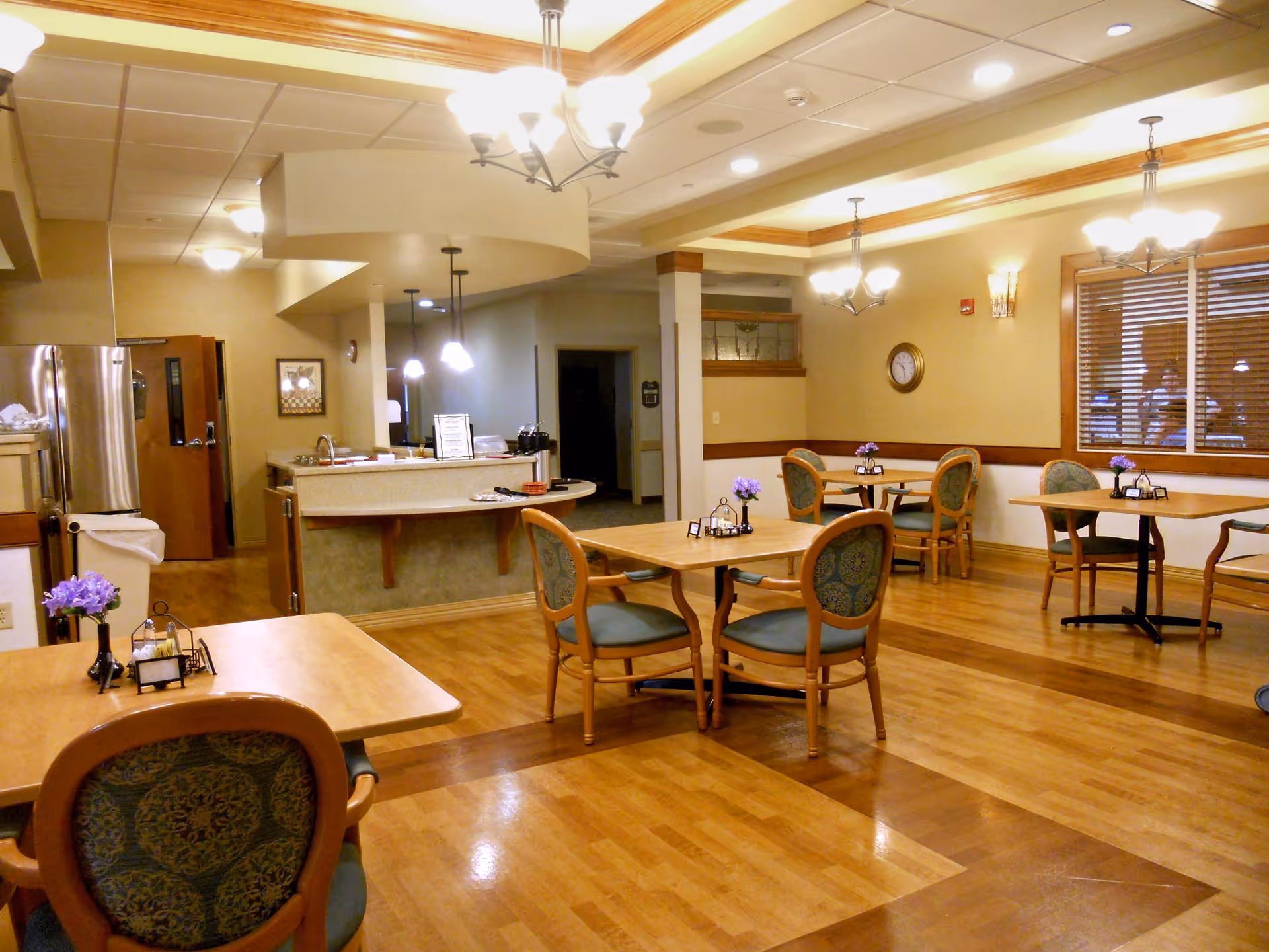 A well-lit dining area in a senior living facility with wooden floors and multiple tables and chairs. Each table has a small vase with purple flowers and condiment holders. The room features warm beige walls, ceiling lights, and a window with wooden blinds. In the background, there is a kitchen area with a refrigerator and a counter.