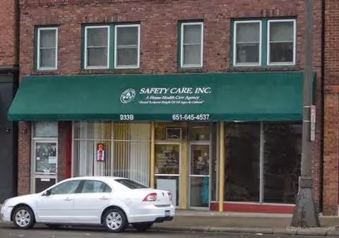 Brick building storefront with green awning reading Safety Care, Inc., and a white car parked outside.