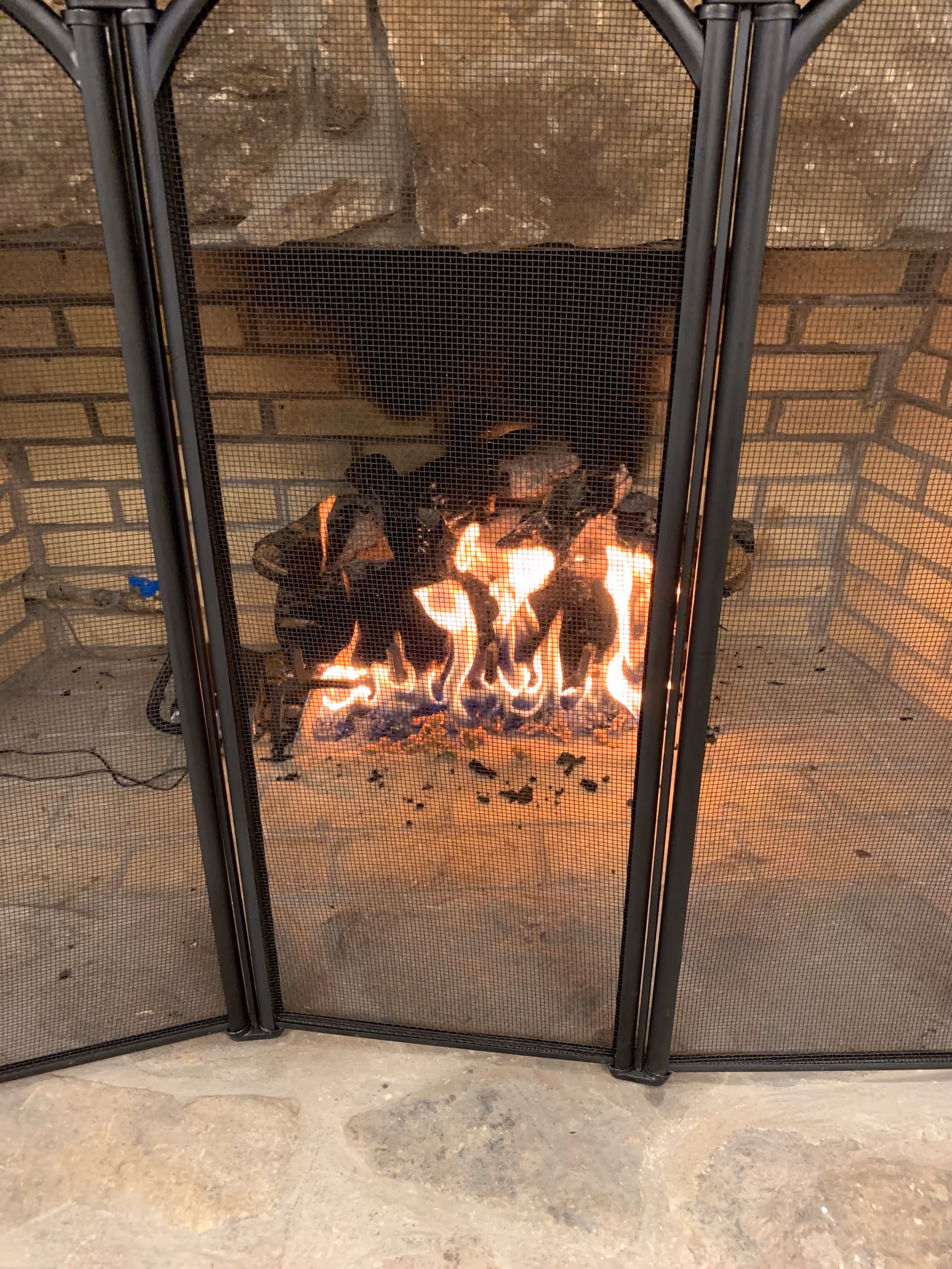 Indoor brick fireplace with glowing flames behind a metal mesh screen.