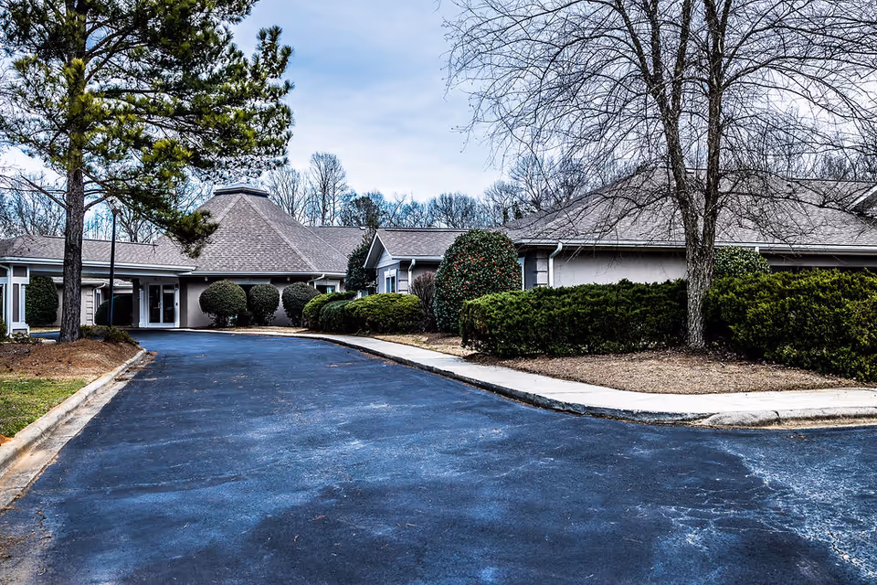 Exterior view of a senior living facility with a paved driveway leading to the entrance. The building has a low, sloped roof and is surrounded by neatly trimmed bushes and trees with no leaves. The sky is overcast.