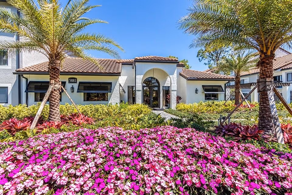 Mediterranean-style building entrance with tiled roof, palm trees, and a bed of bright pink flowers in the foreground.