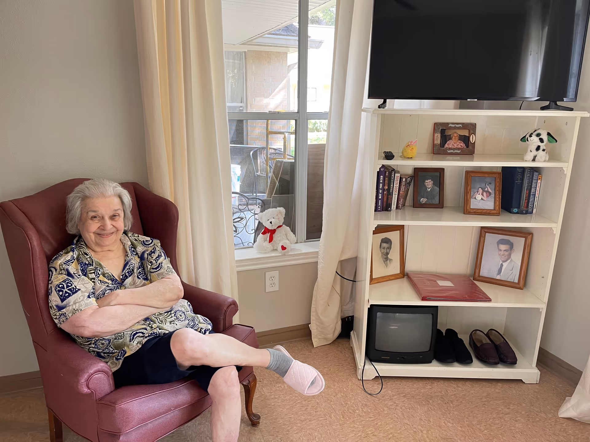 An elderly woman sits smiling in a red armchair beside a window and a white bookshelf holding framed photos, a TV, and personal items.