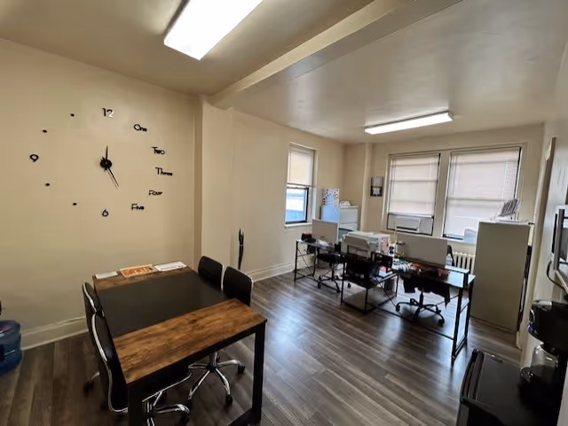 Office-like shared workspace with desks, computers, and a large wall clock in a bright room.