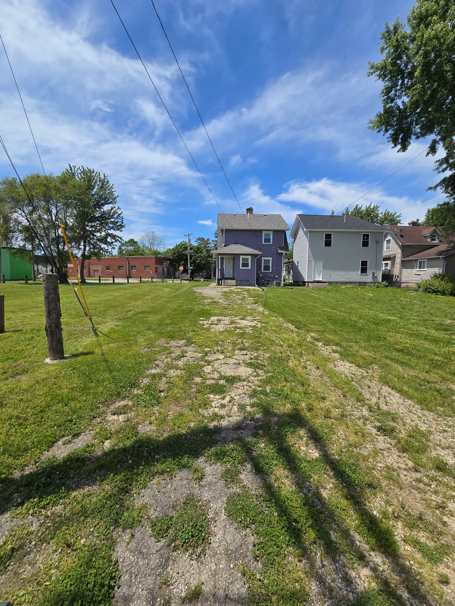 Sunny residential street with grassy front yards, a gravel path, and neighboring houses under blue sky.