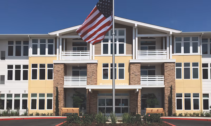 Front view of Merrill Gardens at Huntington Beach, beige building with white trim and flag.