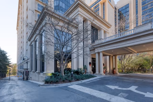 Exterior view of a multi-story building with classical architectural elements including columns and large windows. The entrance is covered by a portico supported by columns, with some greenery and trees around the building. The driveway has directional arrows painted on the pavement.