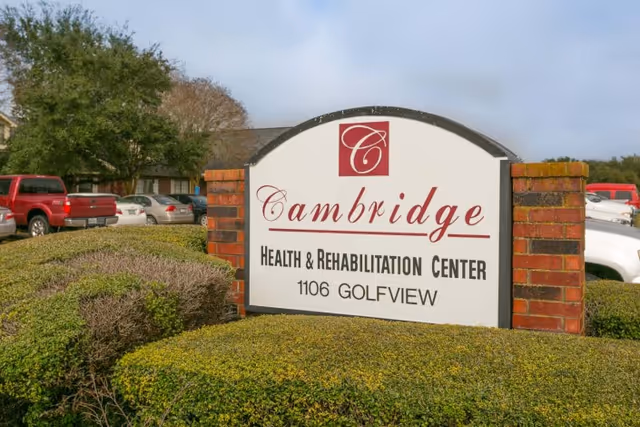 A landscaped front sign reading "Cambridge Health & Rehabilitation Center 1106 GOLFVIEW" with parked cars and bushes behind it.